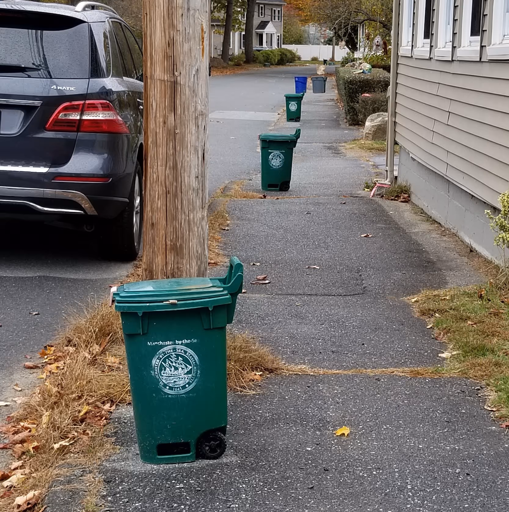 Line of green Manchester-by-the-Sea waste bins along a residential sidewalk next to a parked black SUV.