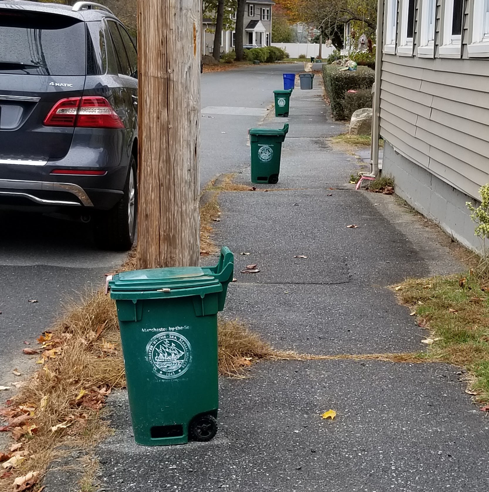 Line of green Manchester-by-the-Sea waste bins along a residential sidewalk next to a parked black SUV.