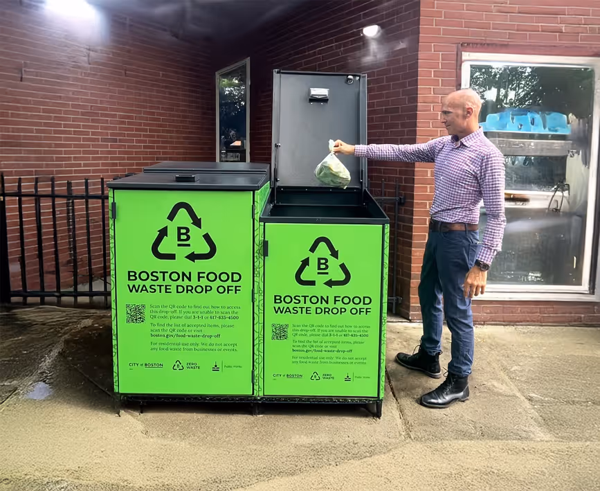 Man in checkered shirt and jeans putting a bag of food waste into a green Boston food waste drop-off bin outside a brick building.