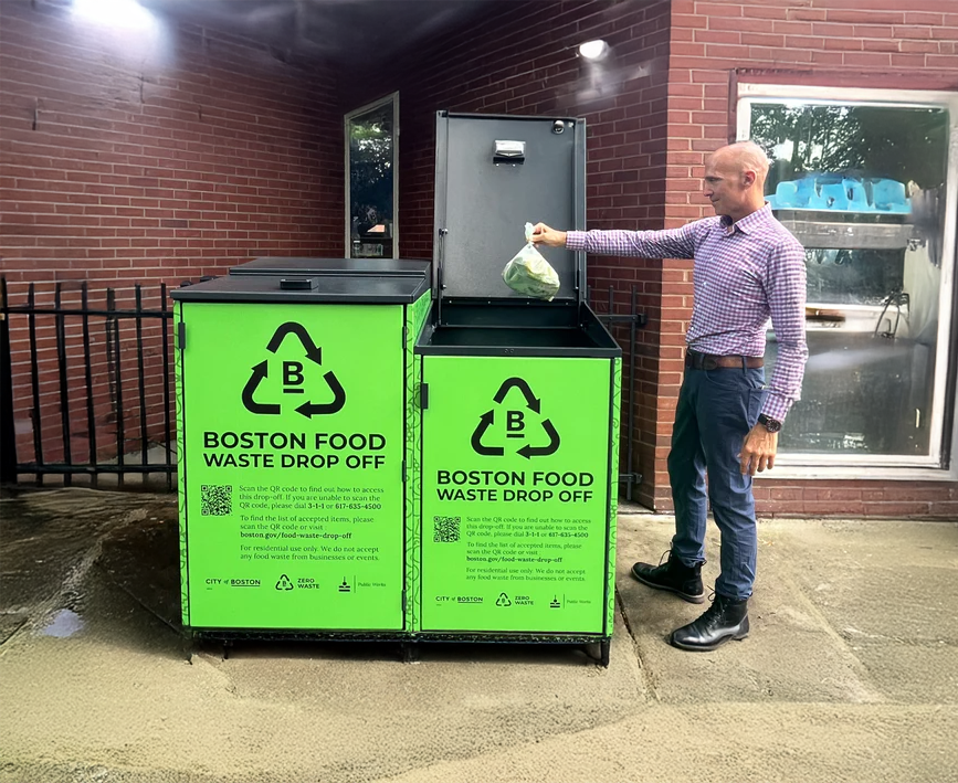 Man in checkered shirt and jeans putting a bag of food waste into a green Boston food waste drop-off bin outside a brick building.