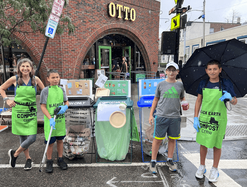 Four people wearing green aprons labeled 'Black Earth Compost crew' stand beside compost, trash, and recycling bins on a rainy street in front of a brick building with 'OTTO' sign.