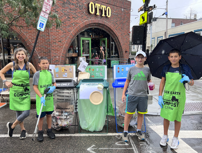 Four people wearing green aprons labeled 'Black Earth Compost crew' stand beside compost, trash, and recycling bins on a rainy street in front of a brick building with 'OTTO' sign.