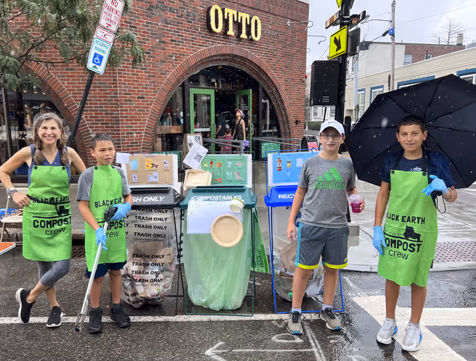 Four people wearing green aprons labeled 'Black Earth Compost crew' stand beside compost, trash, and recycling bins on a rainy street in front of a brick building with 'OTTO' sign.