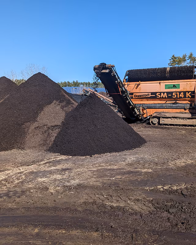 Industrial compost screening machine creating a large pile of dark compost soil outdoors under clear blue sky.