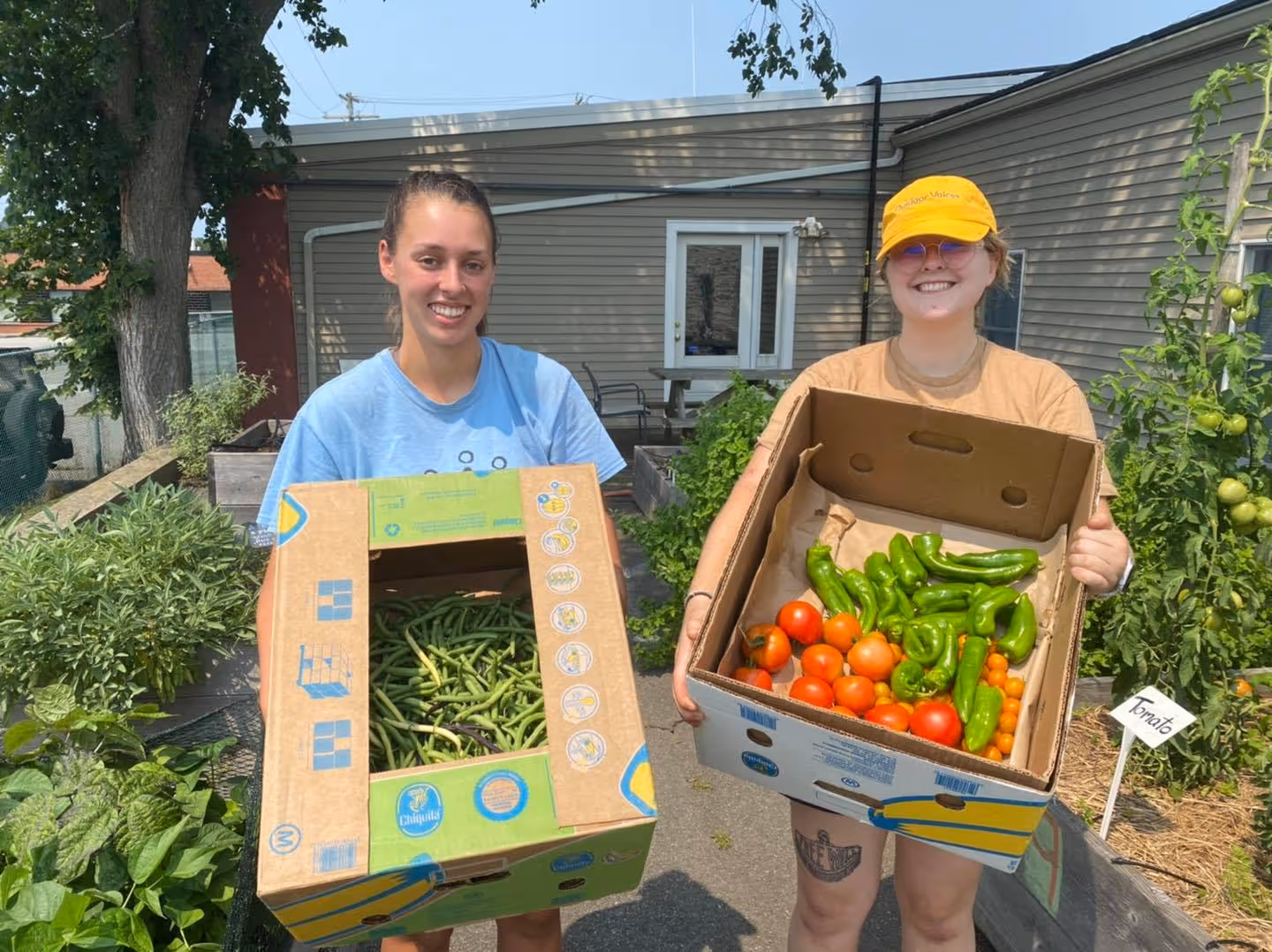 Two smiling women outdoors holding boxes of fresh harvested vegetables including green beans, tomatoes, and green peppers.