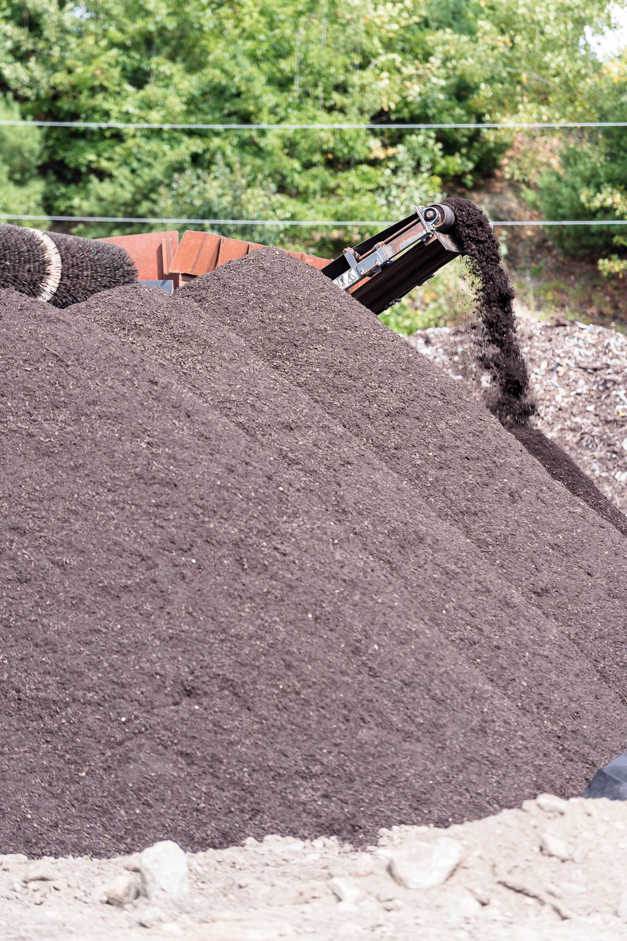 Conveyor belt pouring a large pile of dark mulch with surrounding piles and green trees in the background.