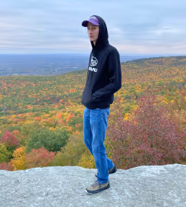 Young man in a black hoodie and cap standing on a rock overlooking a colorful autumn forest landscape.