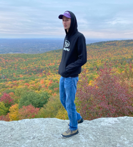 Young man in a black hoodie and cap standing on a rock overlooking a colorful autumn forest landscape.