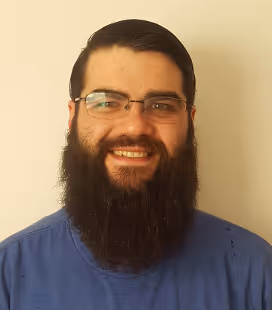 Smiling man with dark thick beard, glasses, and a blue shirt standing against a plain beige wall.