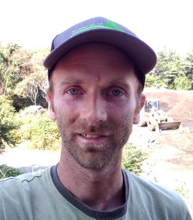 Man in a gray cap and greenish-gray shirt standing outdoors at a construction site with trees and equipment in the background.