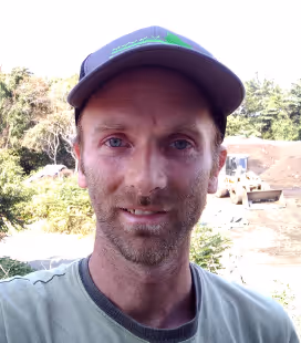 Man in a gray cap and greenish-gray shirt standing outdoors at a construction site with trees and equipment in the background.