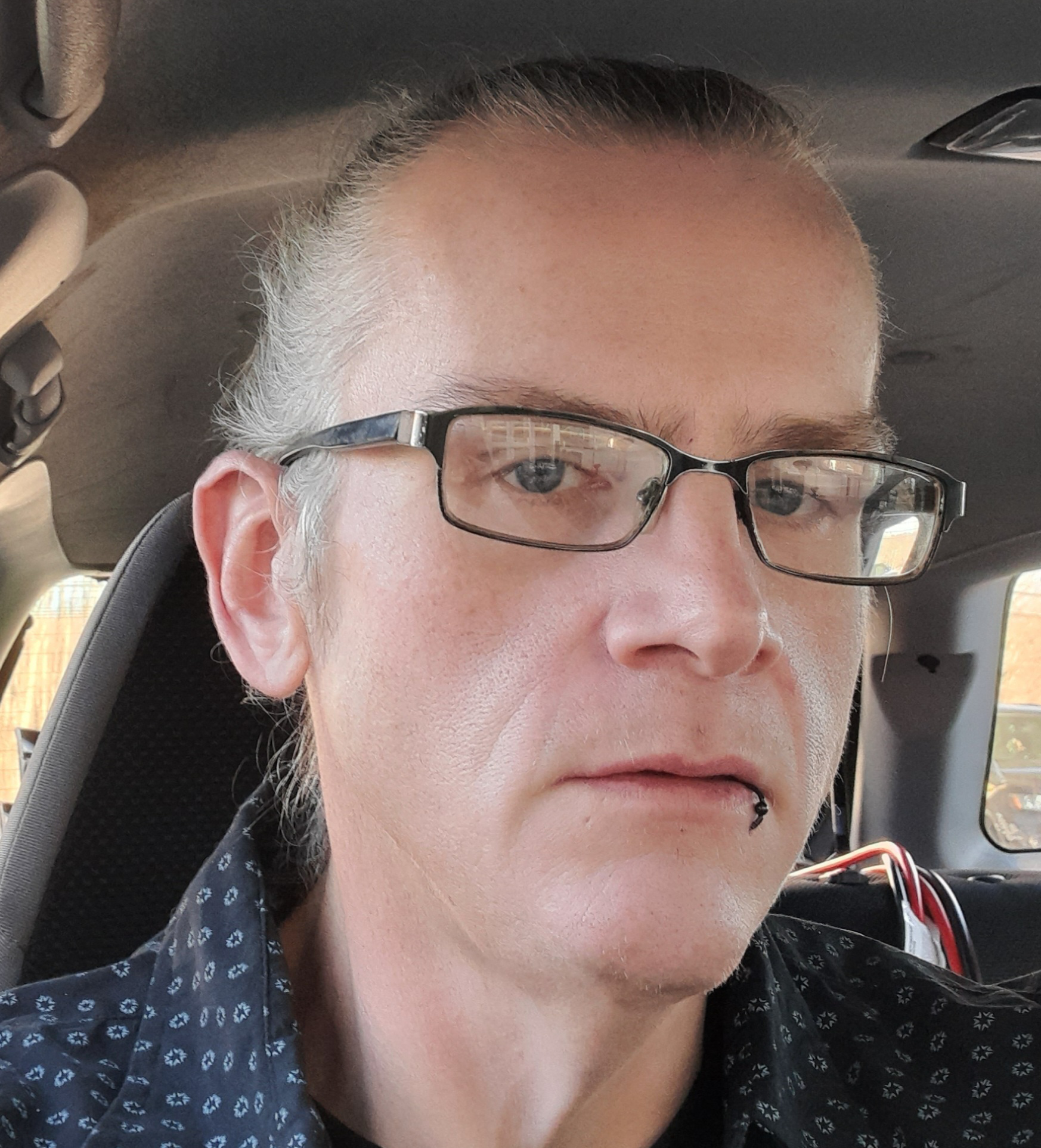 Close-up of a middle-aged person wearing glasses with a lip ring and a patterned shirt, sitting inside a car.