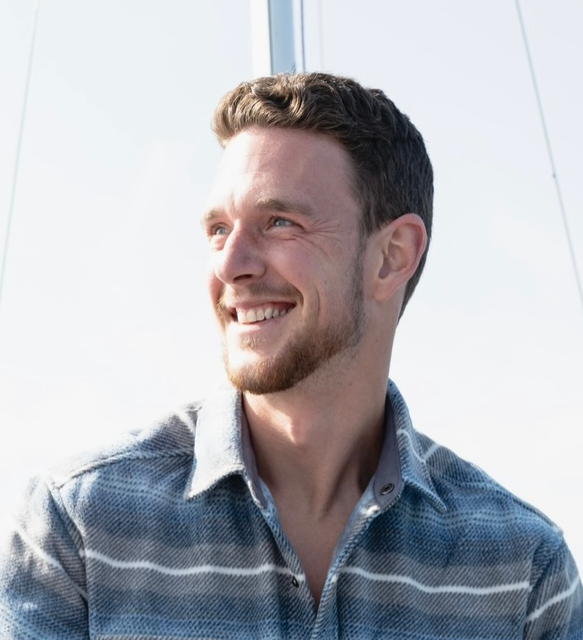 Smiling man with short brown hair and beard wearing a blue plaid shirt outdoors.