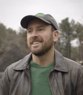 Smiling bearded man in a green sweater and brown jacket wearing a baseball cap outdoors with trees in the background.