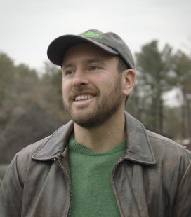 Smiling bearded man in a green sweater and brown jacket wearing a baseball cap outdoors with trees in the background.