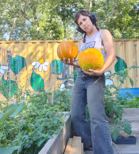 Person standing in a garden holding two large pumpkins, with raised garden beds and a colorful mural in the background.