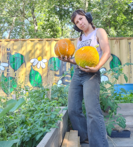 Person standing in a garden holding two large pumpkins, with raised garden beds and a colorful mural in the background.