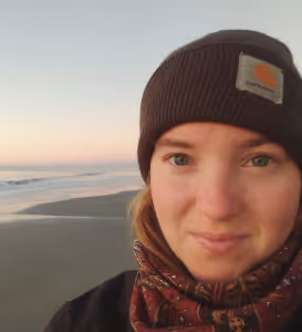 Close-up of a woman wearing a dark beanie and patterned scarf at a beach during sunset.