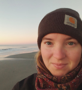 Close-up of a woman wearing a dark beanie and patterned scarf at a beach during sunset.