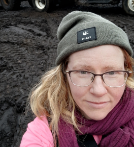 Woman wearing glasses, a green Tilley beanie, a purple scarf, and a pink jacket standing outdoors on muddy ground.