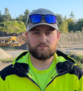 Man with a beard wearing a gray cap, reflective sunglasses, and a neon yellow safety jacket standing outdoors with construction equipment and trees in the background.