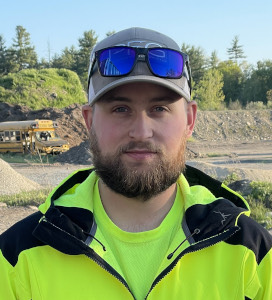 Man with a beard wearing a gray cap, reflective sunglasses, and a neon yellow safety jacket standing outdoors with construction equipment and trees in the background.