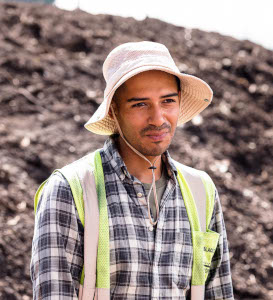 Man wearing a wide-brimmed hat and plaid shirt standing outdoors with soil or dirt mound in the background.