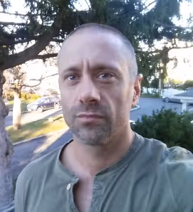 Man with short hair and a trimmed beard wearing a green shirt standing outdoors in front of trees and parked cars.