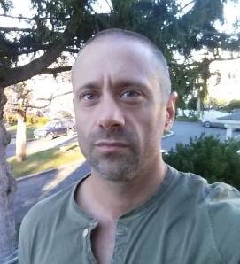 Man with short hair and a trimmed beard wearing a green shirt standing outdoors in front of trees and parked cars.