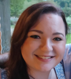 Smiling woman with long brown hair, wearing a blue patterned top, outdoors with greenery in the background.