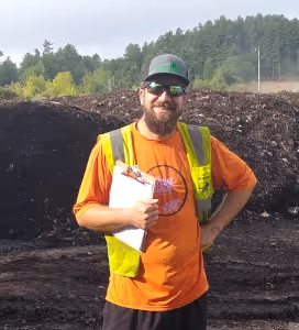 Man wearing an orange shirt and yellow safety vest holding a clipboard standing in front of a large compost or soil pile outdoors with trees in the background.