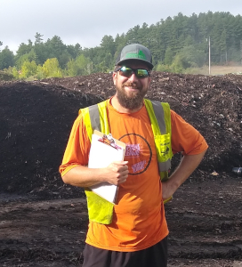 Man wearing an orange shirt and yellow safety vest holding a clipboard standing in front of a large compost or soil pile outdoors with trees in the background.