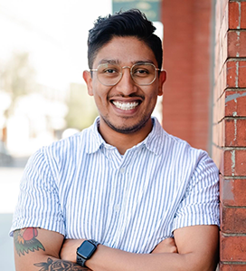 Smiling young man with glasses and tattoos, wearing a striped short-sleeve shirt, standing with arms crossed against a red brick wall.