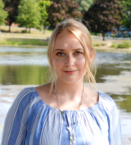 Young woman with blonde hair wearing a blue and white striped blouse standing outdoors near a pond.