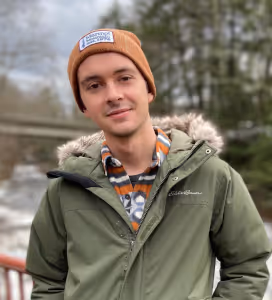 Young man wearing a brown beanie and green winter jacket standing outdoors near a wooden railing with trees and a river in the background.