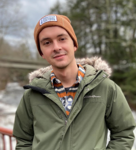 Young man wearing a brown beanie and green winter jacket standing outdoors near a wooden railing with trees and a river in the background.