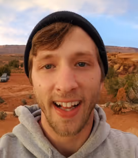 Smiling young man wearing a black beanie and gray hoodie with a desert landscape in the background.