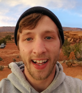 Smiling young man wearing a black beanie and gray hoodie with a desert landscape in the background.