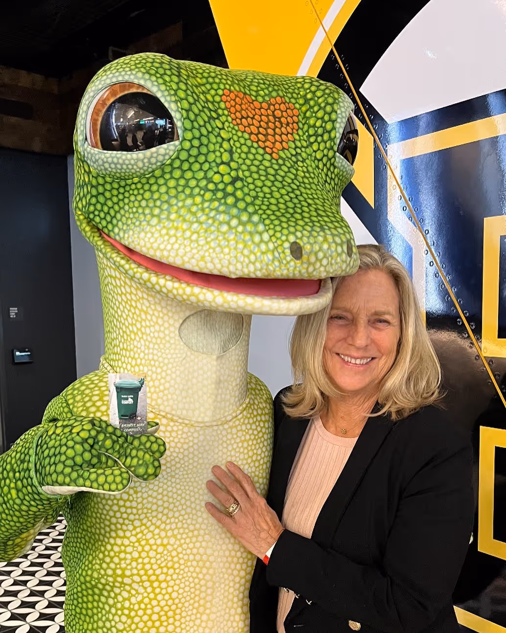 Smiling woman posing with a large green gecko mascot featuring a red heart pattern on its forehead.
