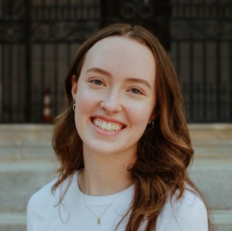 Smiling woman with long wavy brown hair wearing a white top and a small pendant necklace, posing outdoors with steps and a gate in the background.