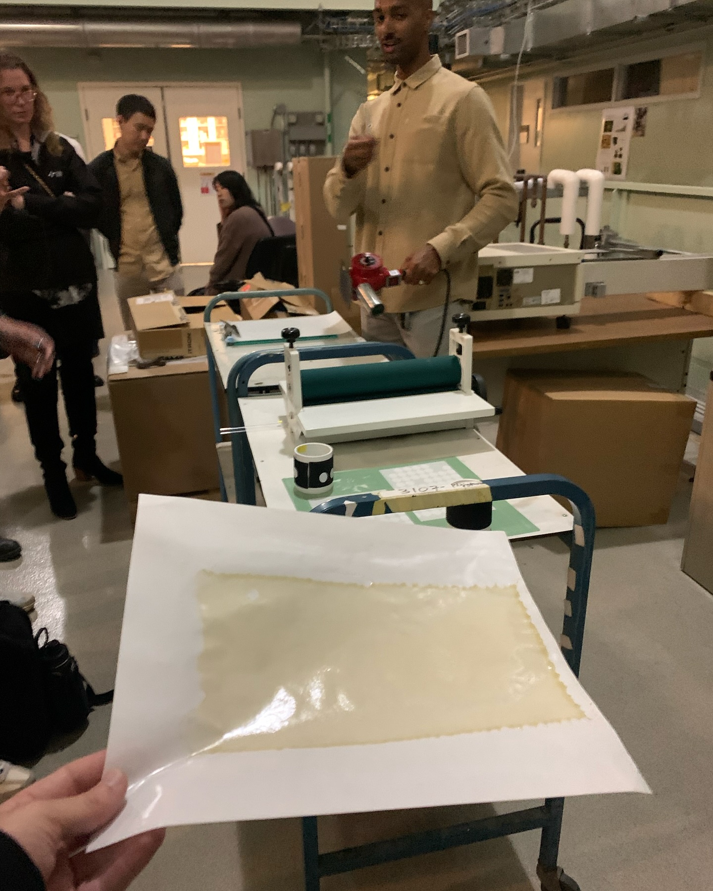 Person holding a sheet of translucent, possibly adhesive material with a machine and people standing in the background in an industrial or workshop setting.