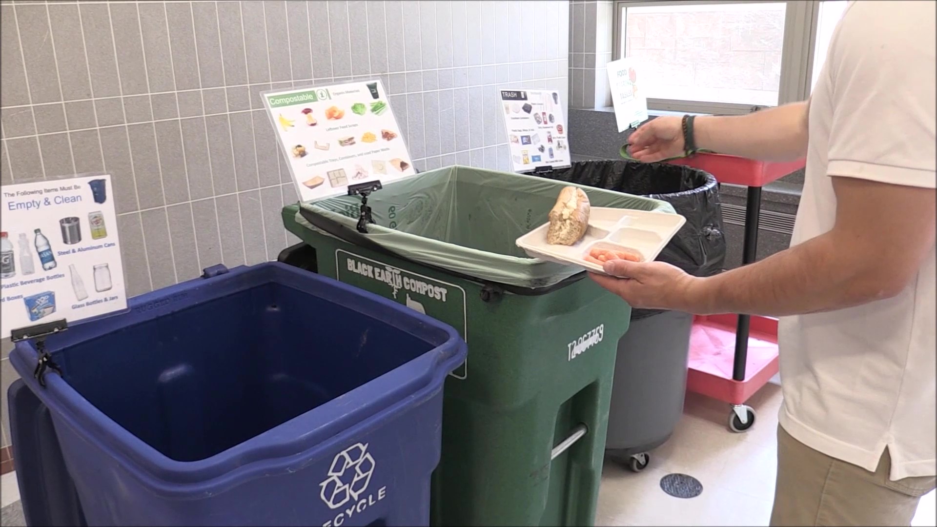 Person holding a food tray with bread and shrimp over a green compost bin next to a blue recycling bin and a black trash bin with sorting signs.