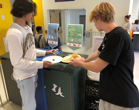 Two teenage boys sorting food waste into a compost bin with a banana peel symbol, guided by signs on proper waste separation in a school cafeteria.