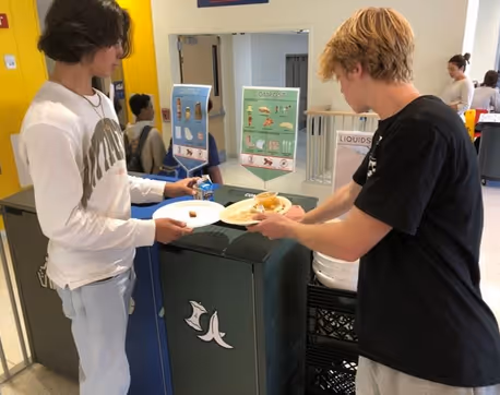 Two teenage boys sorting food waste into a compost bin with a banana peel symbol, guided by signs on proper waste separation in a school cafeteria.