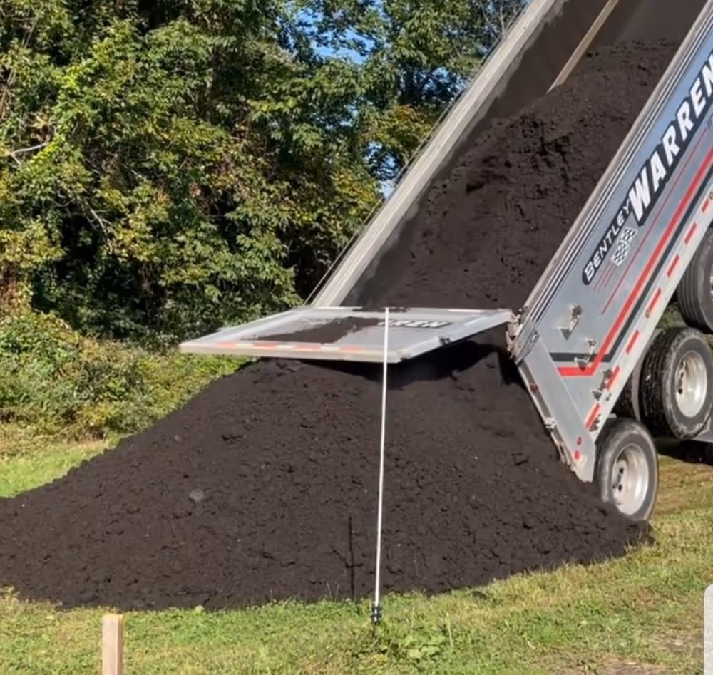Dump truck unloading a large pile of dark soil or compost onto grass near green trees.