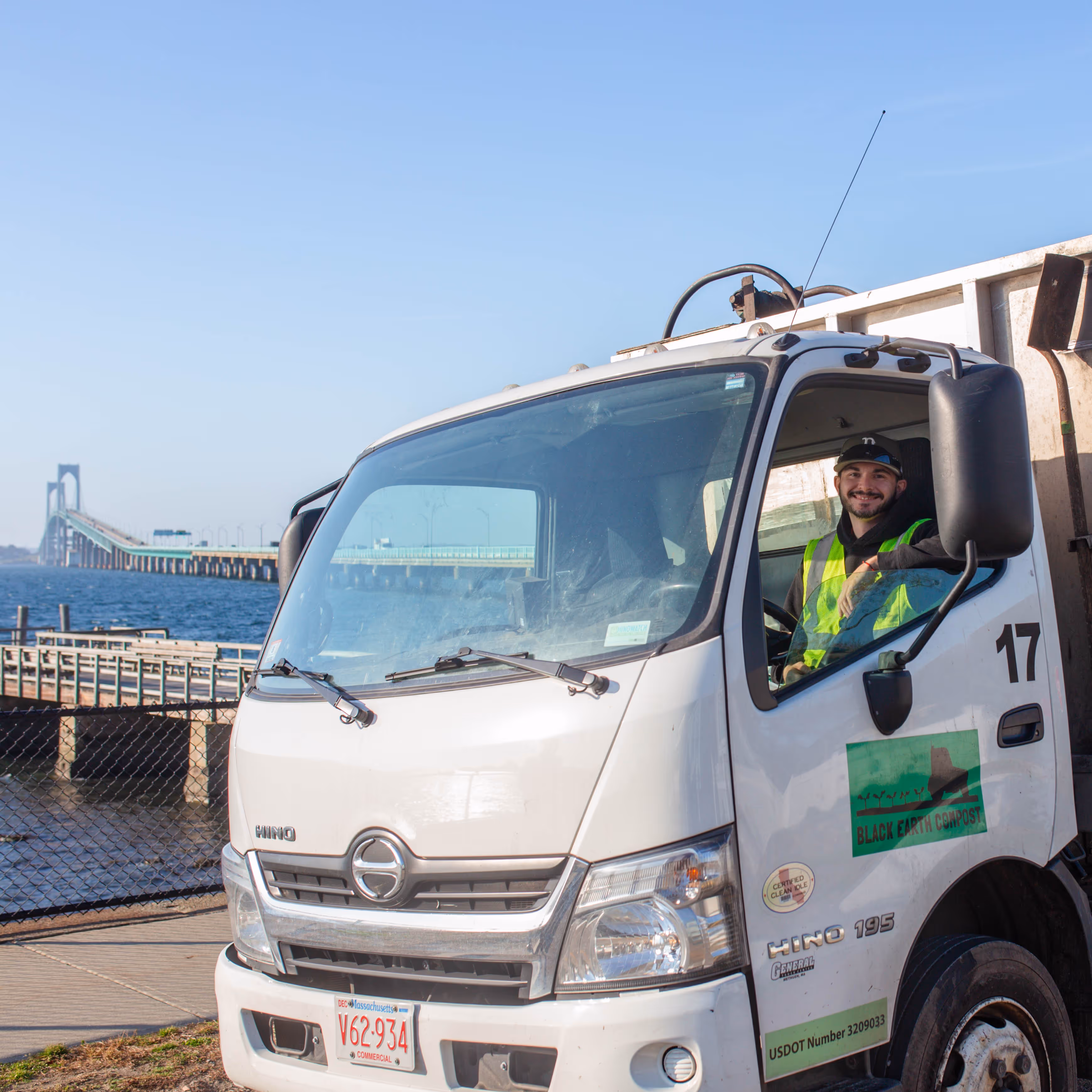 Smiling worker in a yellow safety vest sitting in the driver's seat of a white Black Earth Compost truck near a waterfront with a long bridge in the background.