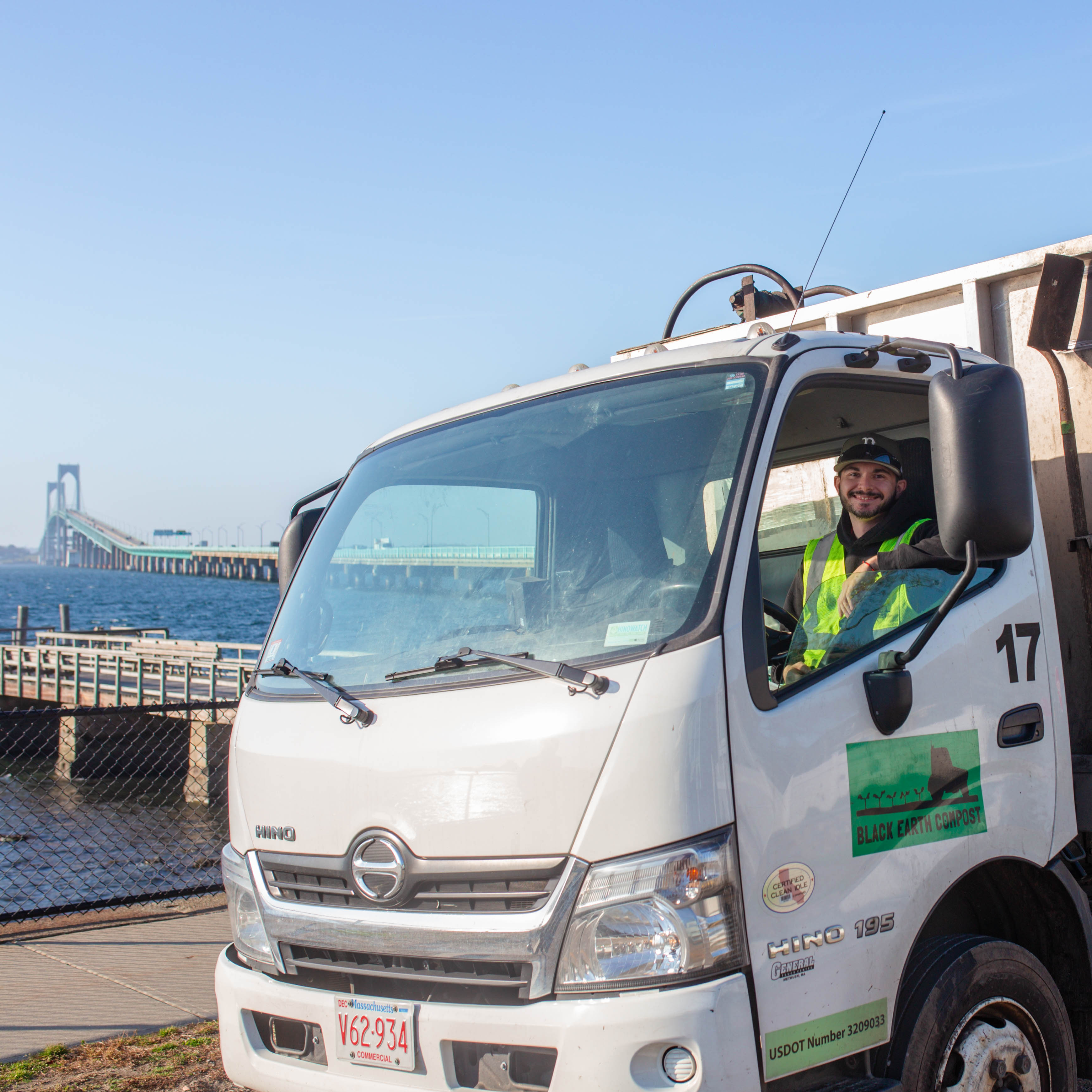 Smiling worker in a yellow safety vest sitting in the driver's seat of a white Black Earth Compost truck near a waterfront with a long bridge in the background.