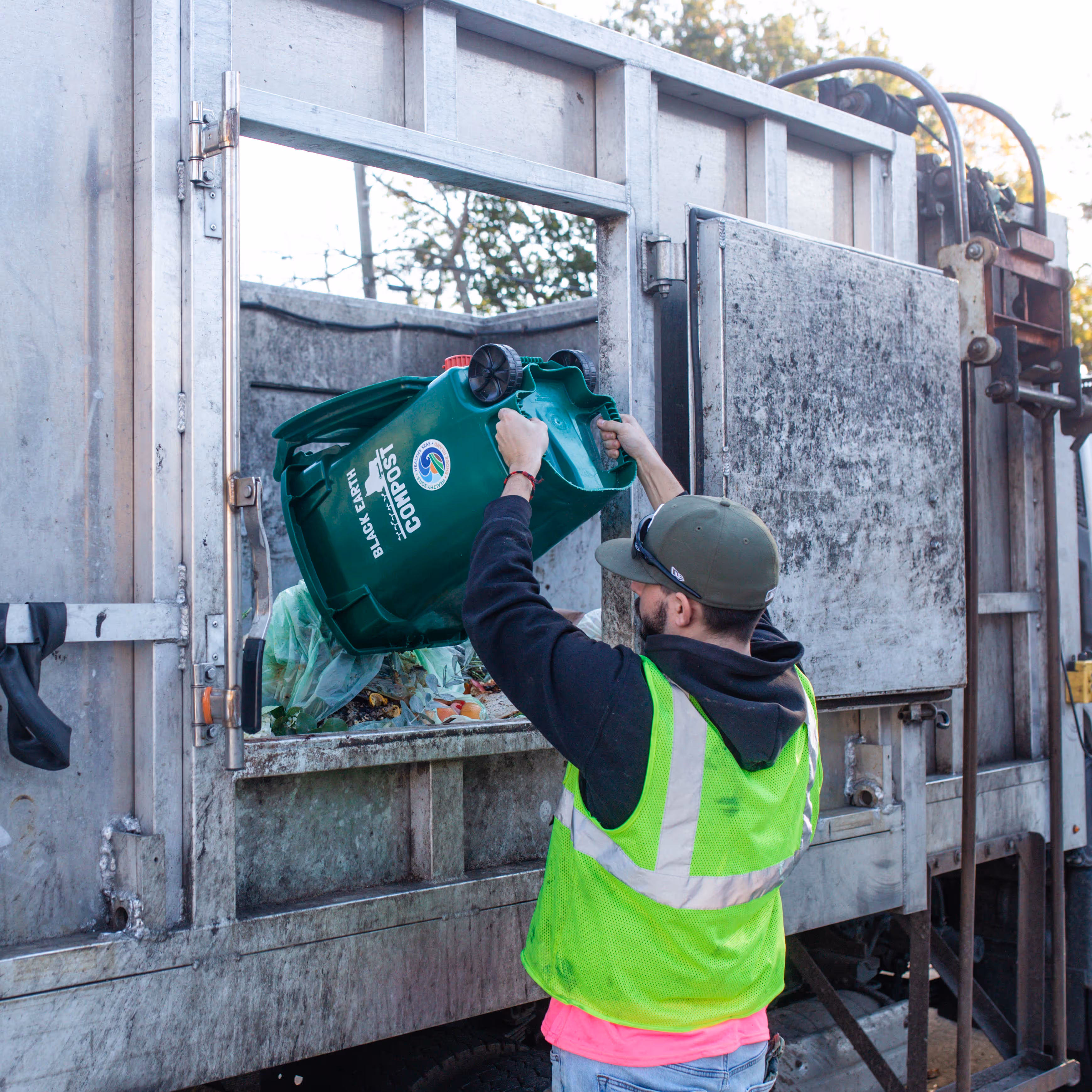 Worker in a neon safety vest emptying a green compost bin into a garbage truck.