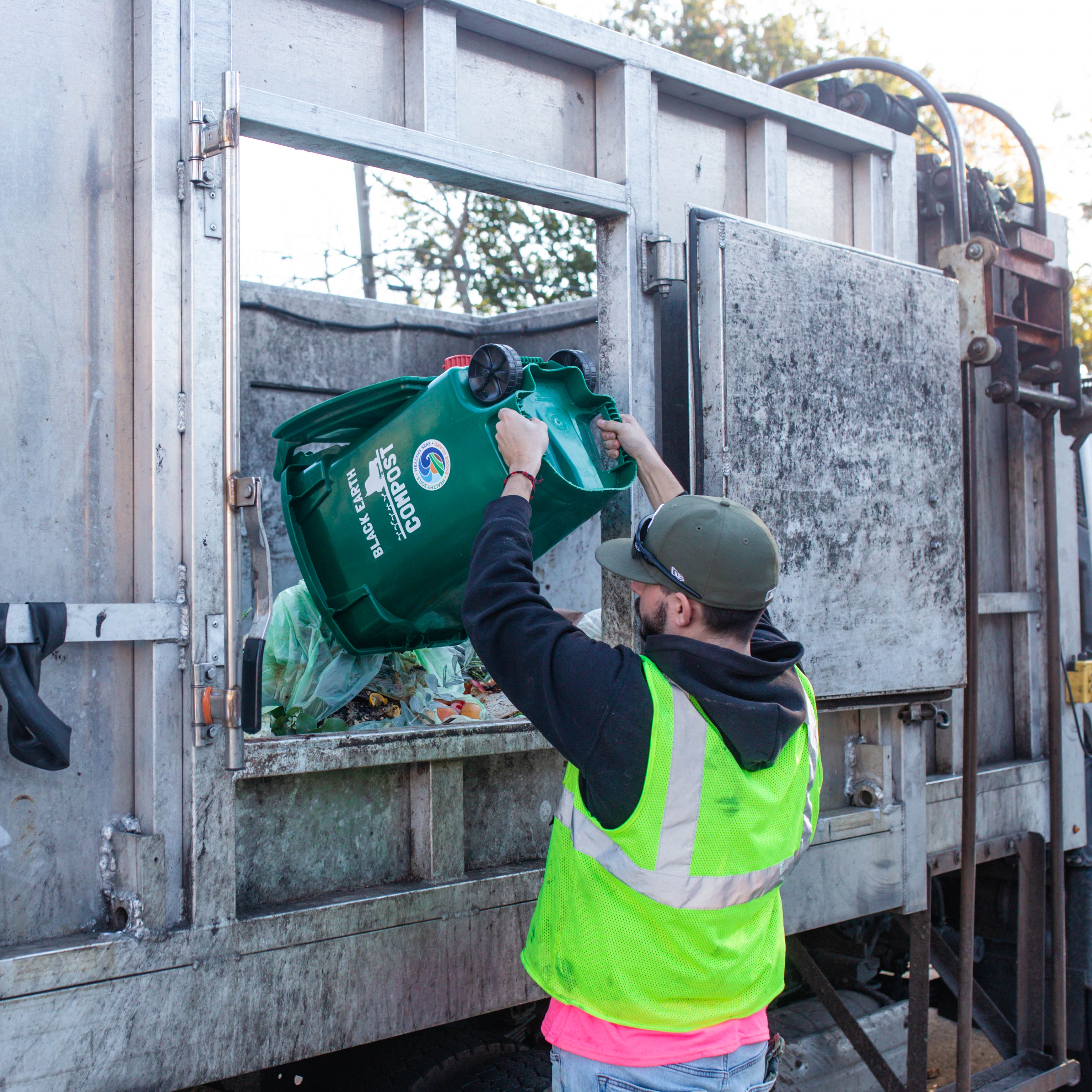 Worker in a neon safety vest emptying a green compost bin into a garbage truck.