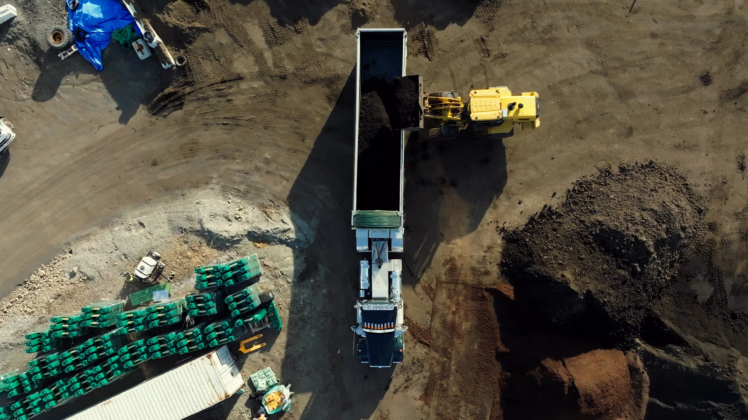 Aerial view of a yellow loader dumping soil into a dump truck at a construction site with green pallets and piles of soil nearby.
